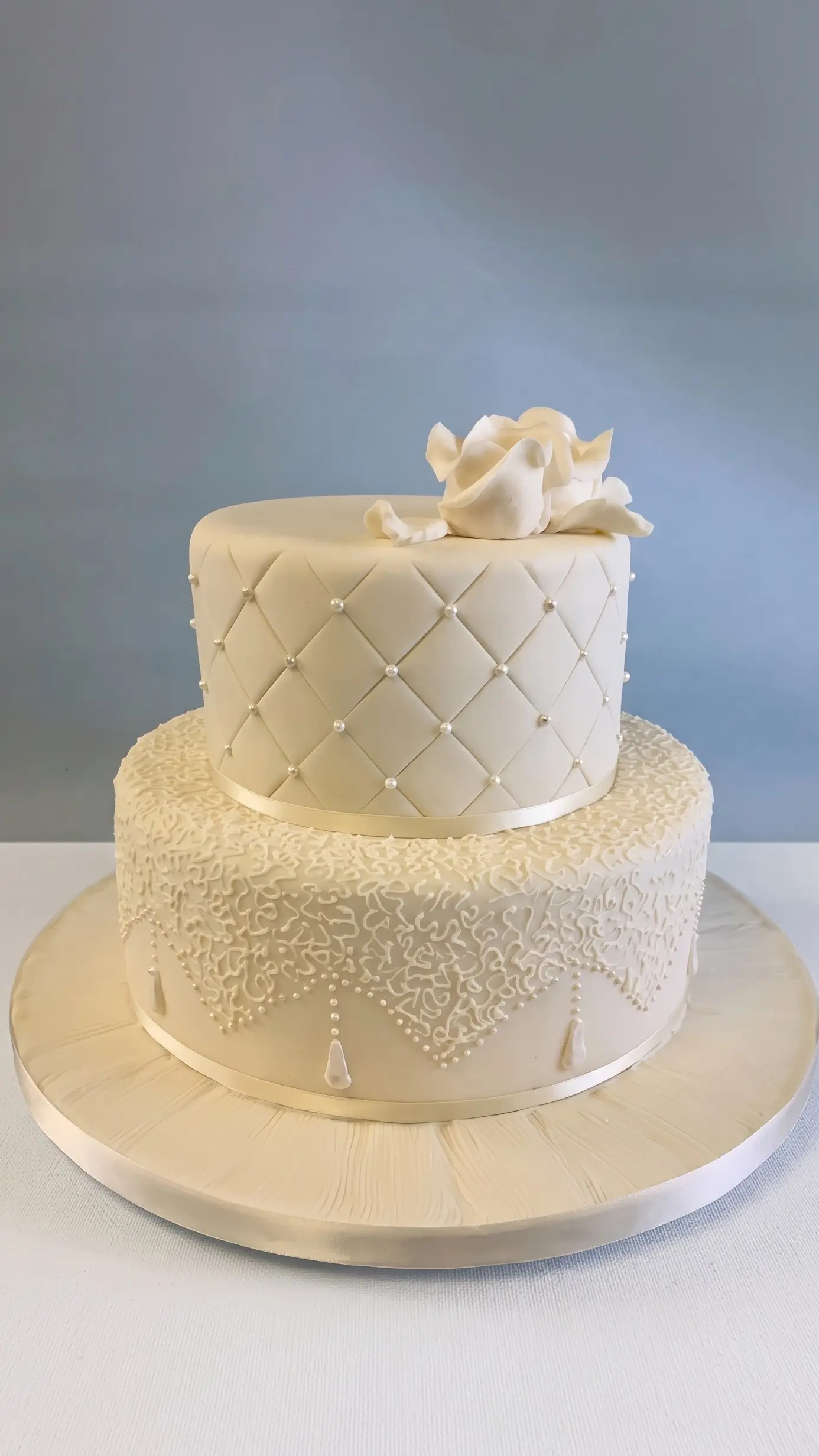 Ivory two-tier wedding cake with quilted pearl details, lace-inspired decoration and white sugar flower topper